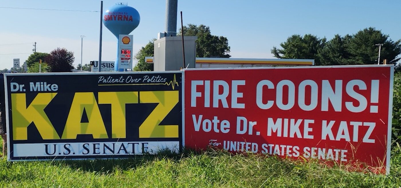 Both Dr. Mike Katz 4-foot by 8-foot signs installed side by side along a Delaware road: the Classic Blue & Gold on the left, the Fire Coons red & white on the right